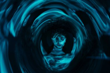 curly man looking at camera through futuristic neon blue circle on dark background 