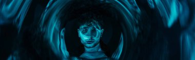 curly man looking at camera through futuristic neon blue circle on dark background, banner