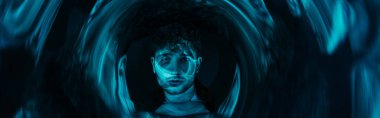 Kıvırcık adam fütüristik neon mavi çemberden kameraya bakıyor.
