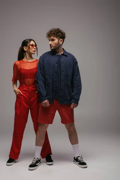 full length of stylish man and woman in trendy outfits posing on grey