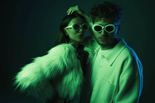 stylish man and woman in sunglasses and trendy outfits posing on dark blue