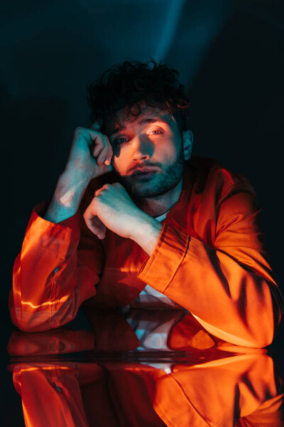 lighting on face of curly and stylish man in orange shirt looking at camera while posing on blue