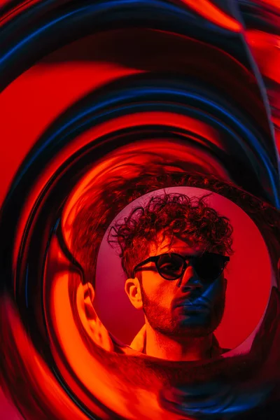 Güneş gözlüklü kıvırcık adam kırmızıdaki gelecekteki neon çemberin ötesine bakıyor.