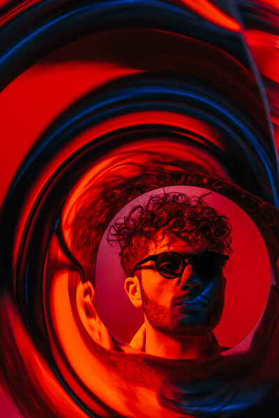 curly man in sunglasses looking away through futuristic neon circle on red