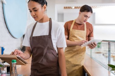 African american seller using digital tablet near blurred colleague in confectionery 