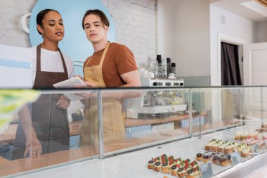 Interracial sellers using digital tablet near eclairs in showcase in sweet shop 