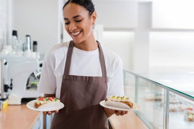 Smiling african american saleswoman holding eclairs in sweet shop 