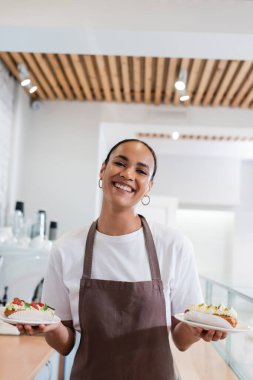 Cheerful african american seller holding eclairs and looking at camera in confectionery