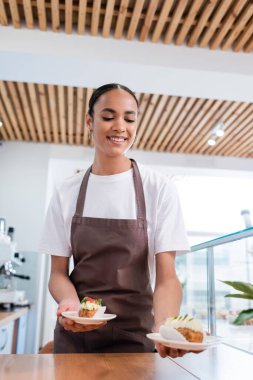 Smiling african american saleswoman holding eclairs in confectionery