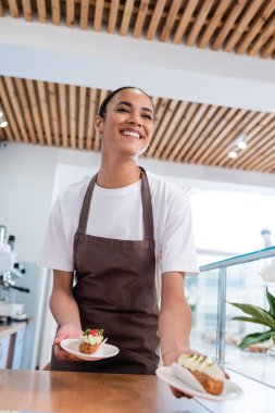 Low angle view of african american seller holding eclairs in confectionery 