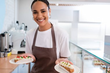 Cheerful african american seller in apron holding eclairs in sweet shop 