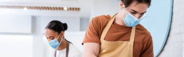 Seller in apron and medical mask working near african american colleague in sweet shop, banner 