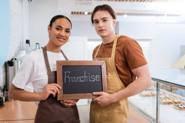 Multiethnic sellers holding chalkboard with franchise lettering in sweet shop  clipart