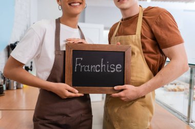 Cropped view of smiling multiethnic sellers holding chalkboard with franchise lettering in confectionery 