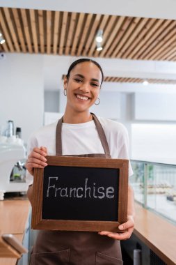 Positive african american seller holding chalkboard with franchise lettering in sweet shop 