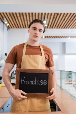 Young seller in apron holding chalkboard with franchise lettering in confectionery 
