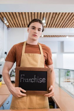 Young seller in apron holding chalkboard with franchise lettering in confectionery  clipart