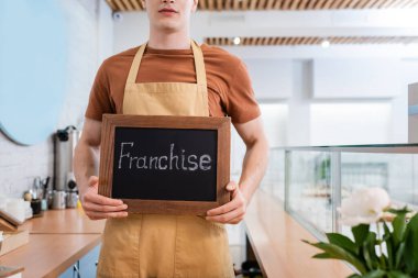 Cropped view of seller holding chalkboard with franchise lettering near showcase and flower in sweet shop 