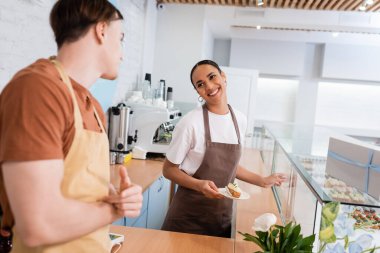 Smiling african american seller holding dessert near showcase and blurred colleague in sweet shop 