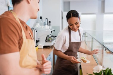 Positive african american seller holding eclair near blurred colleague in confectionery 