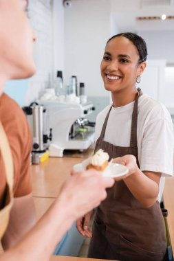 African american seller in apron giving blurred dessert to colleague in confectionery 