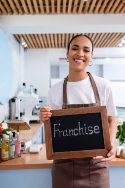 Young african american saleswoman holding chalkboard with franchise lettering in sweet shop  clipart