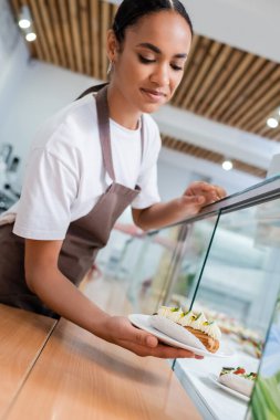 African american saleswoman in apron holding eclair near showcase in confectionery 