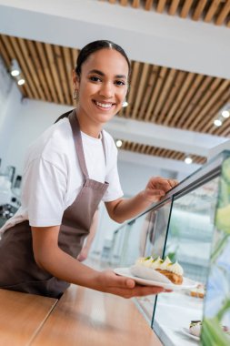 Smiling african american seller holding eclair near showcase in confectionery 