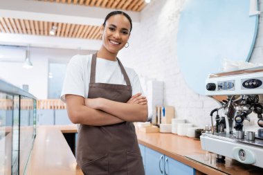 Önlüklü gülümseyen Afro-Amerikan barista şekerci dükkanında kahve makinesinin yanında kollarını kavuşturuyordu. 
