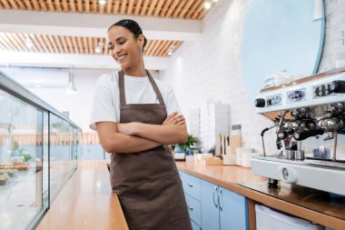 Smiling african american barista looking at showcase near coffee machine in confectionery 