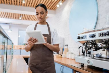 Young african american saleswoman using digital tablet near coffee machine in sweet shop  clipart