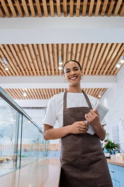 Low angle view of cheerful african american seller holding digital tablet near showcase in confectionery 