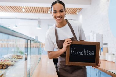Happy african american saleswoman holding chalkboard with franchise lettering near desserts in showcase of sweet shop  clipart