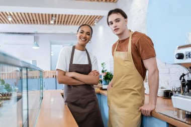 Cheerful multiethnic sellers looking at camera near showcase and coffee machine in sweet shop 