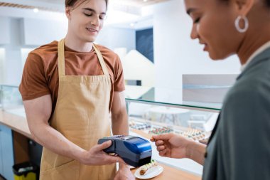 Smiling seller holding payment terminal near african american customer in confectionery 