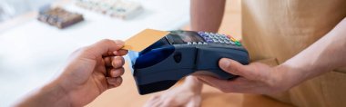 Cropped view of salesman holding terminal near african american customer with credit card in confectionery, banner 