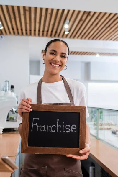 Positive african american seller holding chalkboard with franchise lettering in sweet shop