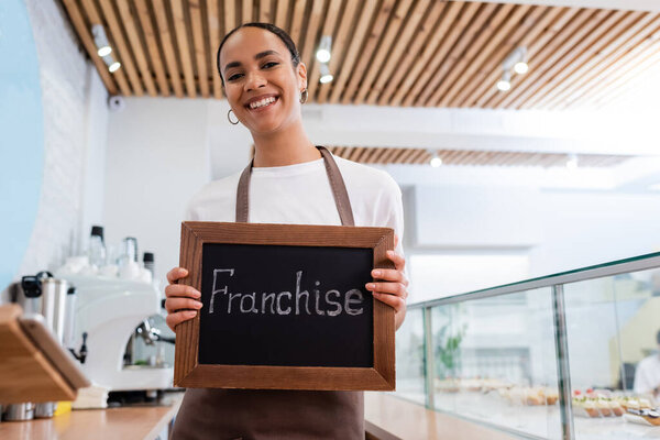 Positive african american seller holding chalkboard with franchise lettering near showcase in sweet shop 