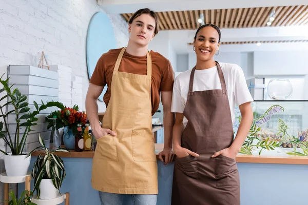 Young Interracial Sellers Aprons Looking Camera Plants Sweet Shop —  Fotos de Stock