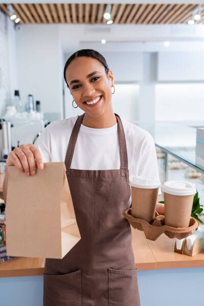 African american seller holding takeaway coffee and paper bag in sweet shop 