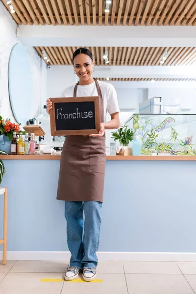Positive african american seller holding chalkboard with franchise lettering in confectionery