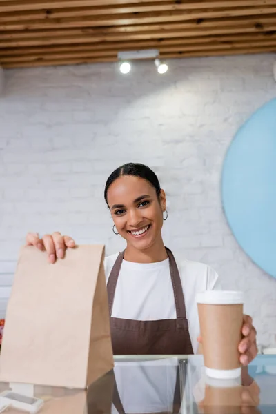 Positive african american seller holding paper cup and bag in sweet shop