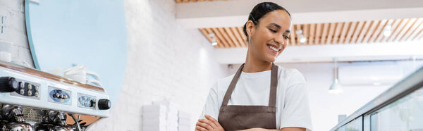 Cheerful african american barista looking at showcase near coffee machine in confectionery, banner 