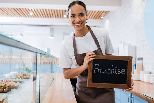 Happy african american saleswoman holding chalkboard with franchise lettering near desserts in showcase of sweet shop 