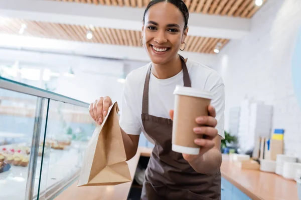 Cheerful african american seller holding paper bag and cup while looking at camera in confectionery