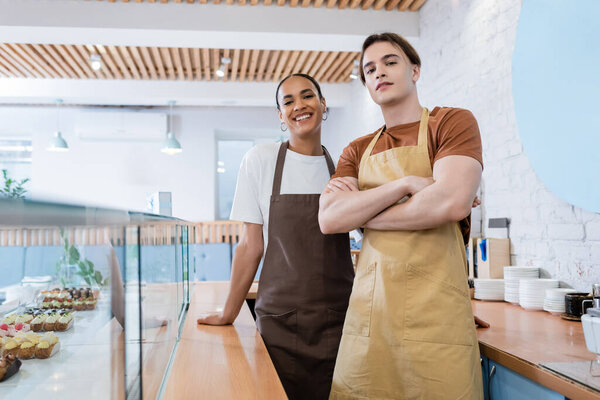 Multiethnic sellers looking at camera near desserts in showcase in confectionery 