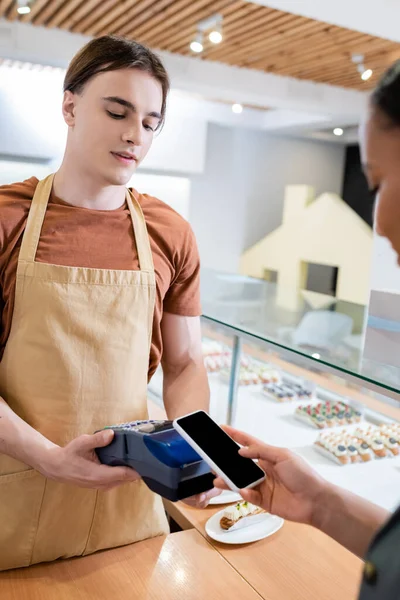 Seller holding payment terminal near african american customer with smartphone in sweet shop