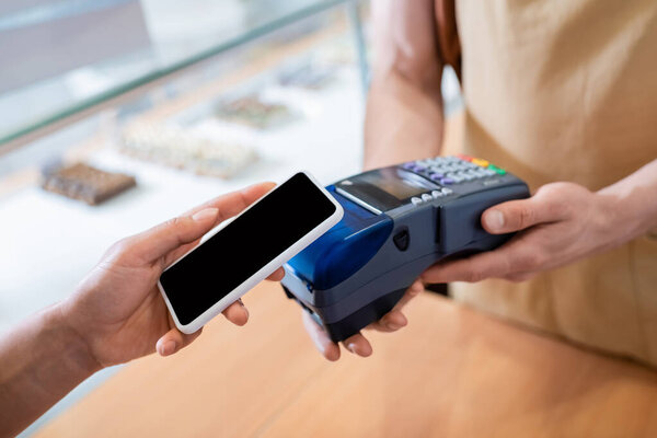 Cropped view of african american woman paying with smartphone near salesman in confectionery 