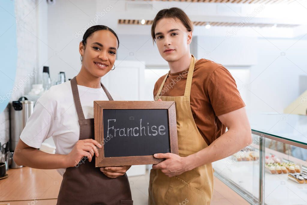 Multiethnic sellers holding chalkboard with franchise lettering in sweet shop 