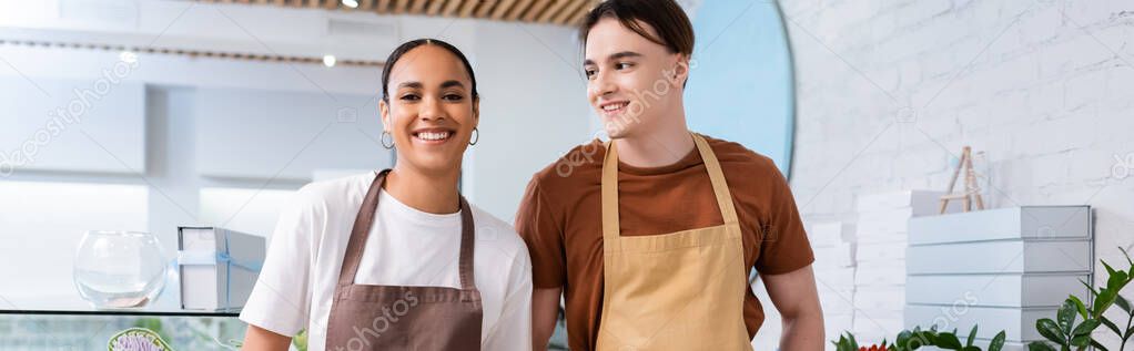 Smiling salesman in apron looking at african american colleague in sweet shop, banner 